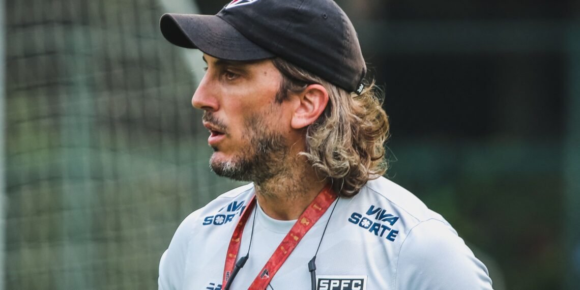 Homem com boné preto e camisa do São Paulo FC no campo, aparentando concentração. Fundo desfocado de rede de futebol.