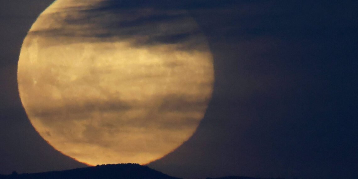 Última superlua de 2025 acontece nesta quinta-feira, 4 de dezembro 31 Lua cheia grande aparece parcialmente encoberta por nuvens finas e escuras, com silhueta de colina ao fundo em céu escuro.