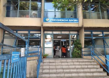 A imagem mostra a entrada de uma agência da Previdência Social. A fachada é de um prédio de dois andares, com janelas grandes e uma escada de acesso. Acima da entrada, há uma placa azul com o texto