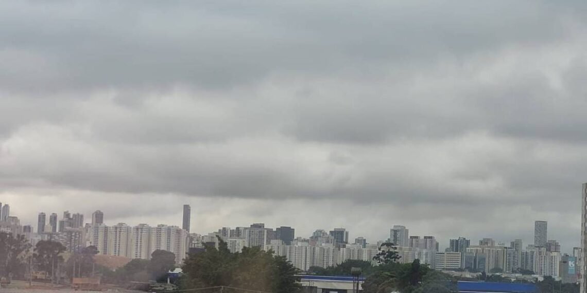 Vista panorâmica da cidade com prédios residenciais e comerciais sob um céu carregado de nuvens cinzentas. Áreas verdes e construções de diferentes alturas compõem a paisagem urbana.