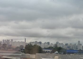 Vista panorâmica da cidade com prédios residenciais e comerciais sob um céu carregado de nuvens cinzentas. Áreas verdes e construções de diferentes alturas compõem a paisagem urbana.