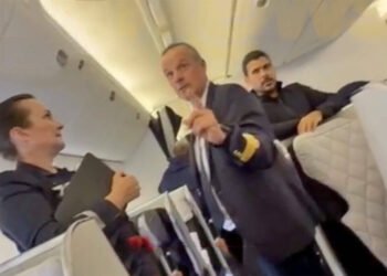 Família da Bahia é retirada de voo por mudança de classe 35 Três comissários de bordo estão no corredor de uma cabine de avião. Dois deles estão em pé, um segurando um tablet e outro apontando para a câmera, enquanto o terceiro está sentado ao fundo. As poltronas são claras e o ambiente é iluminado por luzes embutidas no teto.