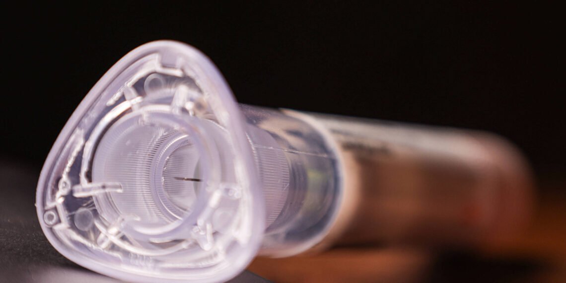 A imagem mostra uma seringa em close, com foco na parte superior que tem uma forma triangular. A seringa é transparente e apresenta detalhes internos visíveis, como um êmbolo e uma agulha. O fundo é desfocado, sugerindo um ambiente escuro.